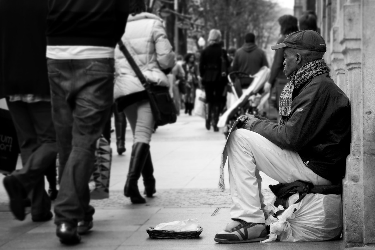 A black-and-white image of a homeless person sitting on the streets holding a cardboard sign and with a small basket in front to collect money, while other people walk past them.